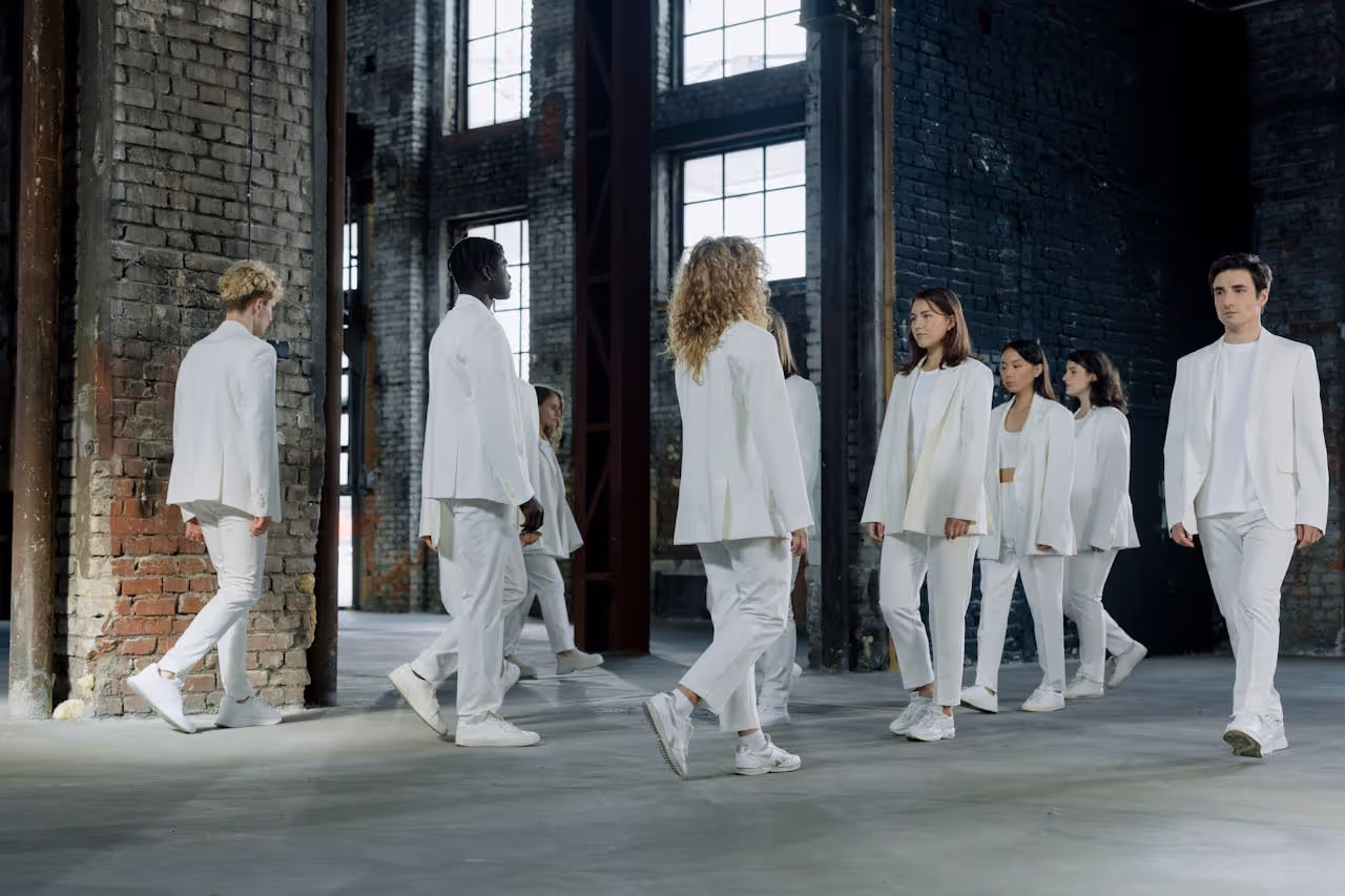 Group of men and women dressed in white suits walking in an industrial-style room with brick walls and large windows.