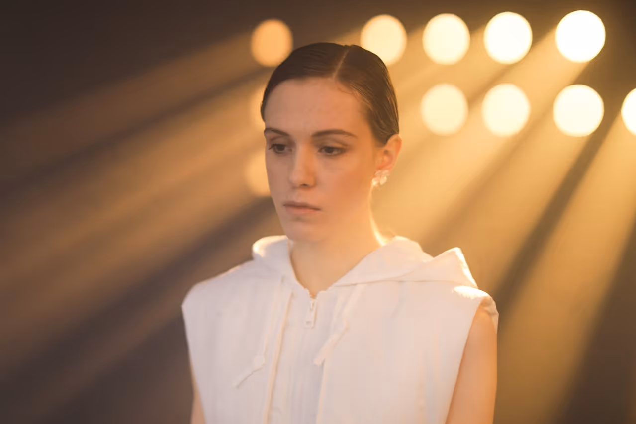 Young woman with slicked-back hair wearing a sleeveless white hoodie under warm, bright stage lights.
