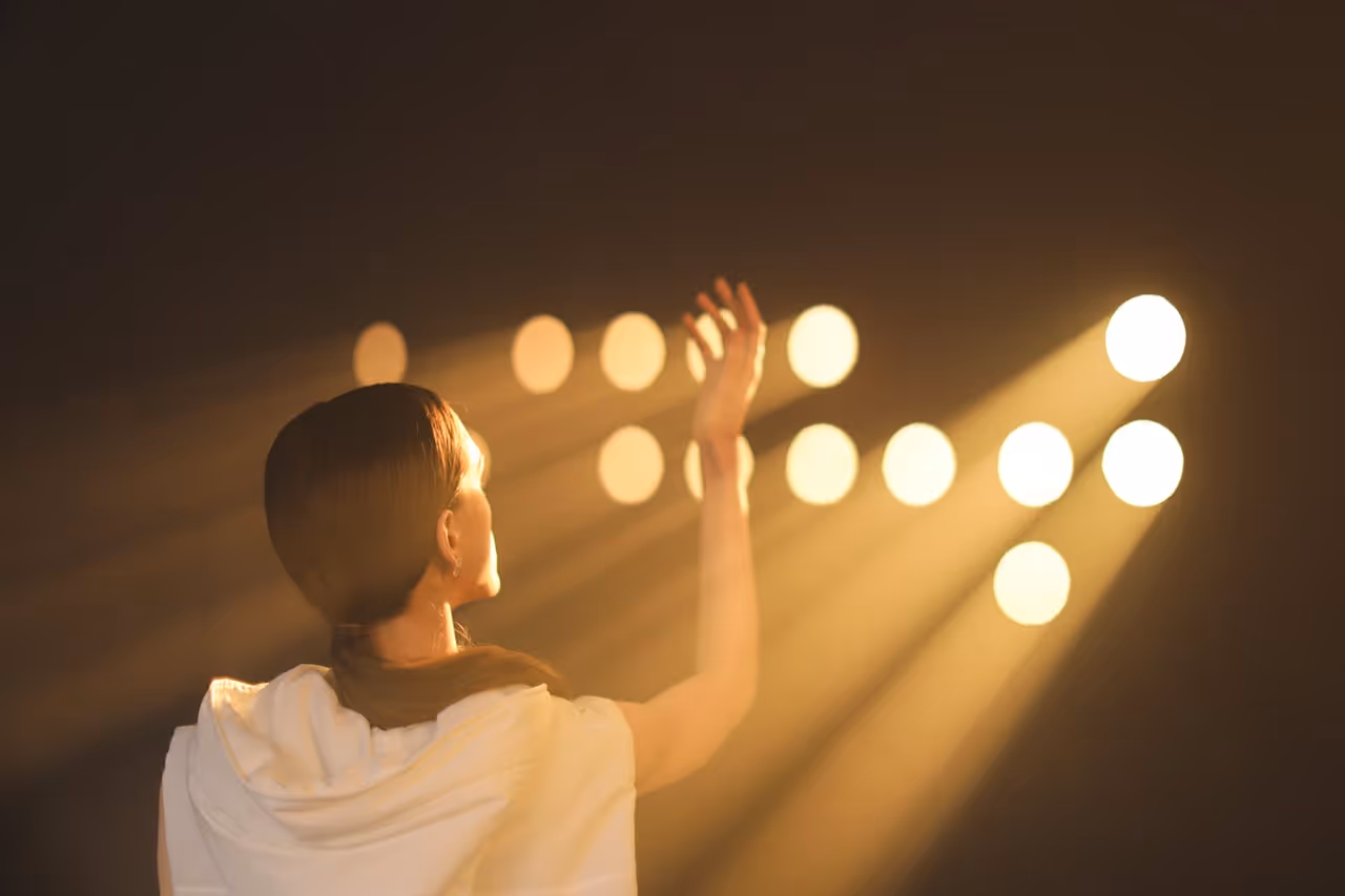 Woman with her hand raised reaching toward bright stage lights in a dark space.