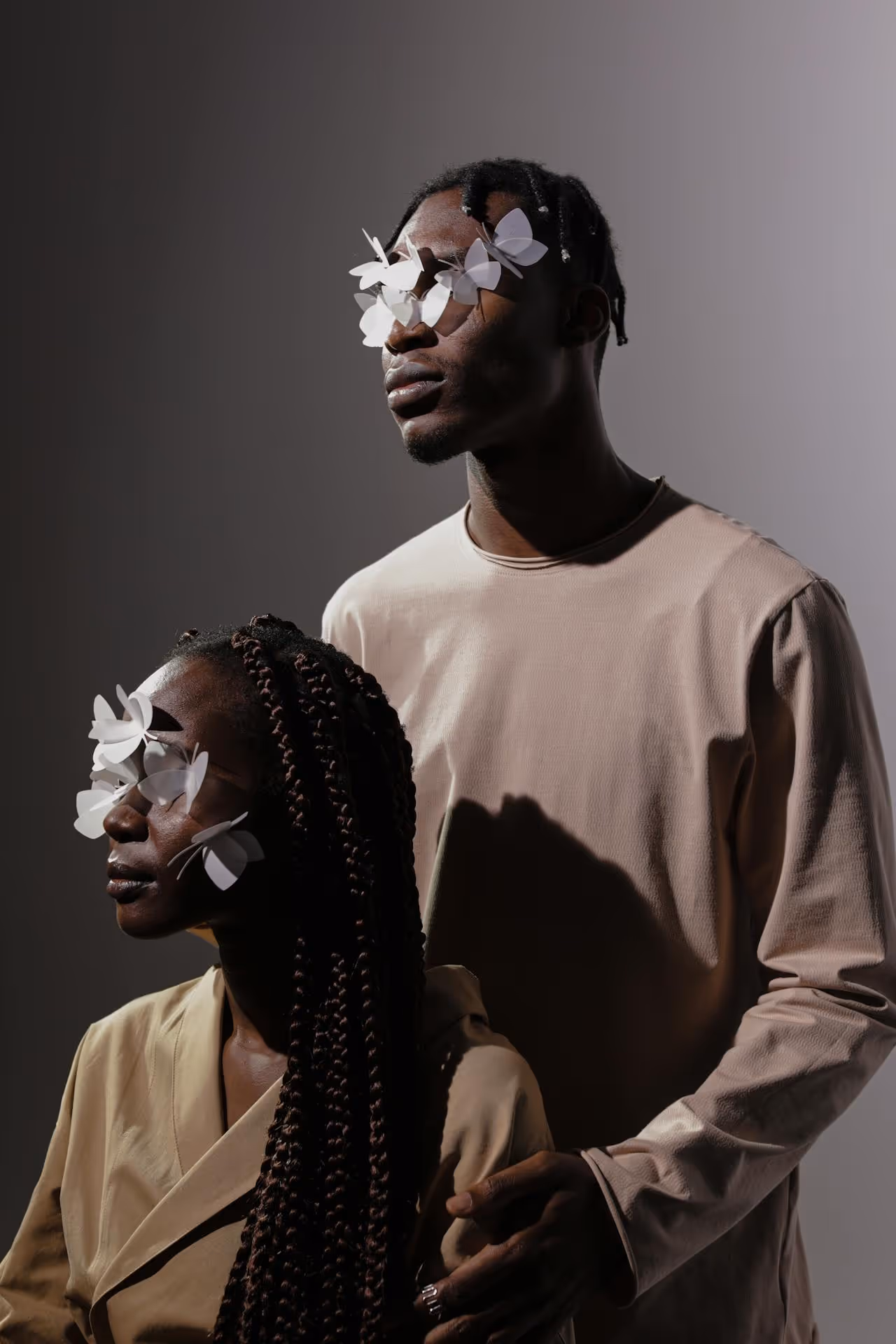 Two people with dark skin and braided hair wearing beige clothing, each with white paper butterflies covering their eyes.