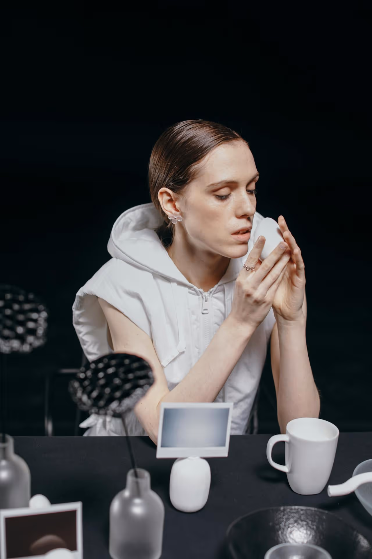 Person in white sleeveless hooded garment holding a white cup close to their face at a table with black decorative objects and a white mug.