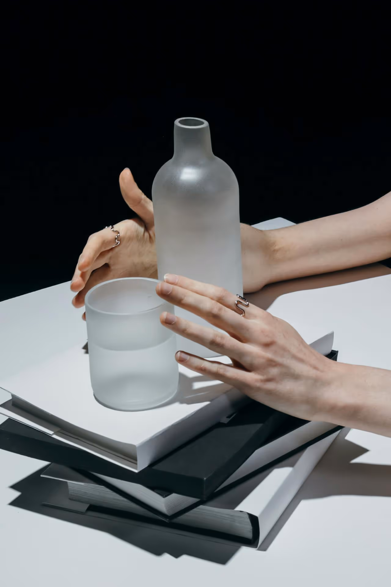 Hands with silver rings holding a frosted glass bottle and drinking glass atop a stack of black and white books on a white table.