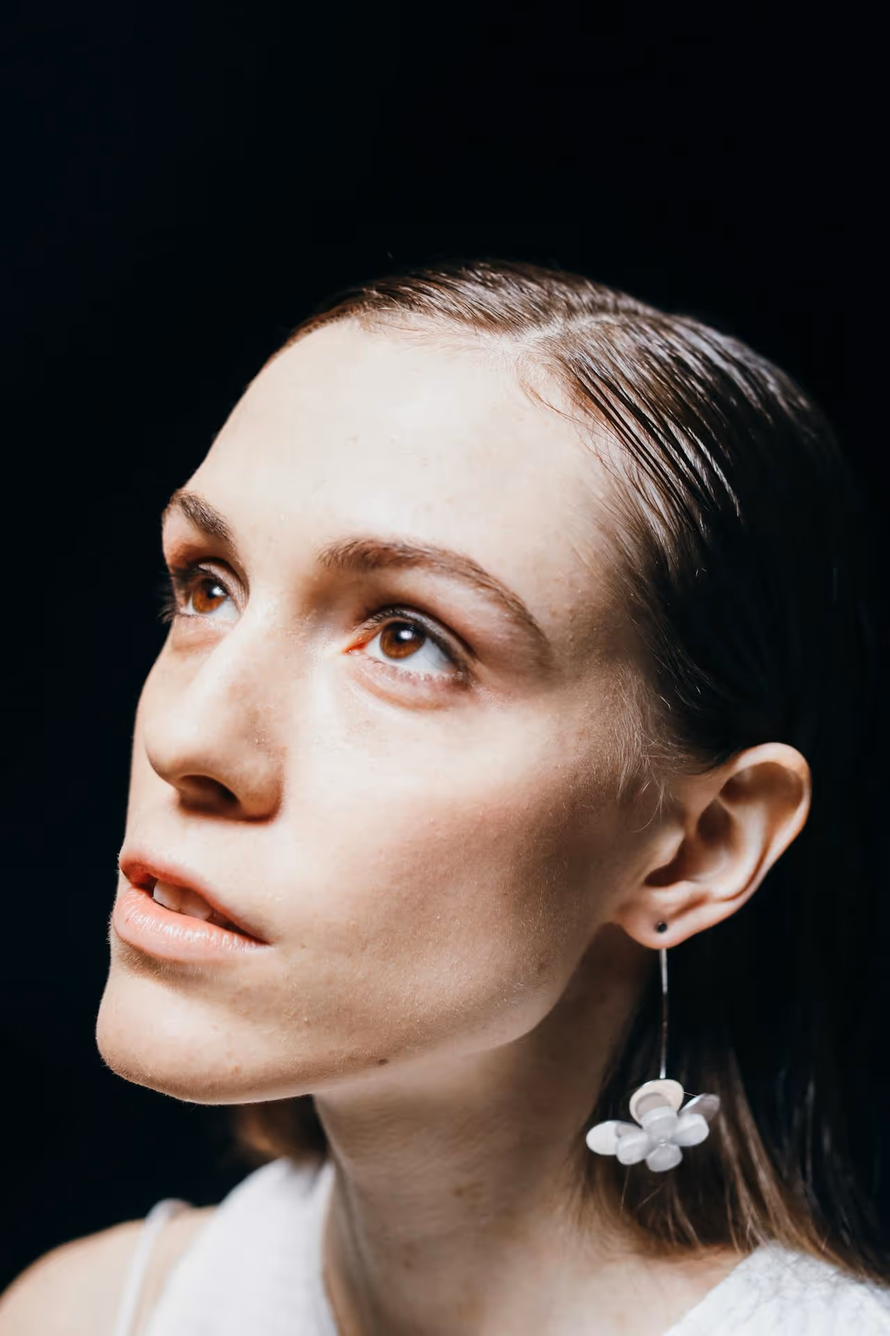 Close-up portrait of a woman with wet hair wearing a white flower-shaped earring looking upward against a black background.