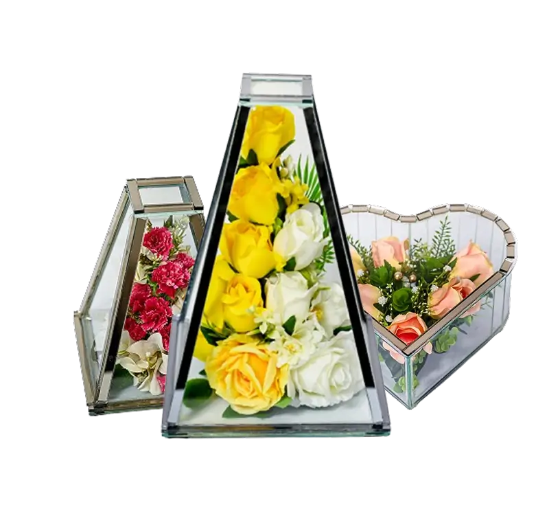 Three glass terrariums containing colorful flower arrangements: yellow and white roses in a pyramid-shaped terrarium, pink carnations in a smaller geometric terrarium, and peach roses in a heart-shaped terrarium.
