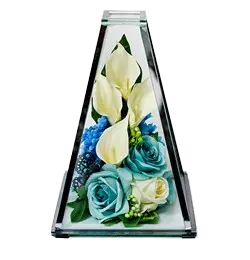 Modern triangular glass vase containing white calla lilies, blue roses, white rose, and small white flowers.
