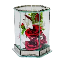 Hexagonal glass display case containing two red roses, white lilies, and green foliage on a mirrored base.