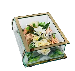Rectangular glass box with gold edges containing a floral arrangement of white, cream, and peach flowers with green leaves.