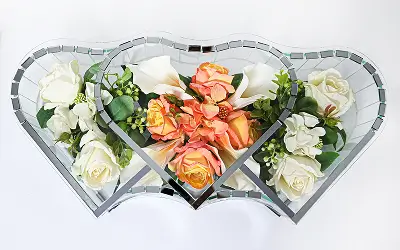 Heart-shaped mirrored glass container holding a bouquet of white and orange roses with green foliage.