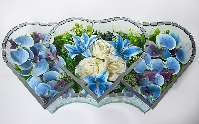 Decorative mirrored heart-shaped frame holding blue orchids, white roses, and green foliage.