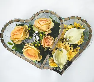 Heart-shaped mirrored tray holding artificial peach and brown roses with green leaves on a white background.