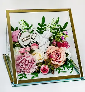 Decorative arrangement of pink and white roses, green leaves, small pink flowers, two white bird figurines, and a small lantern inside a glass and brass frame.