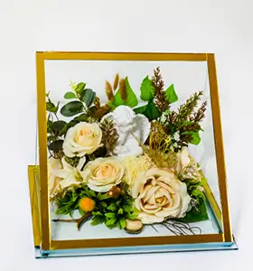 Decorative arrangement of pink and peach roses, white flowers, greenery, and a small angel figurine inside a slanted glass and gold frame.