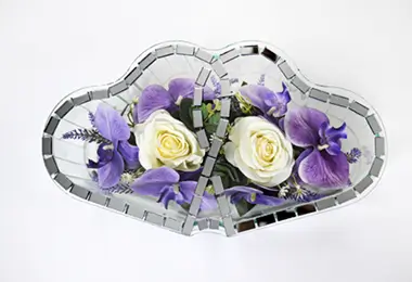 Decorative mirrored glass heart-shaped container holding pink and white artificial flowers with green leaves.