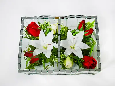 Glass book-shaped vase containing arrangement of white lilies, red roses, green leaves, and white rosebuds on white background.