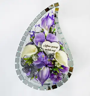 Tear-shaped mirror frame surrounding an arrangement of purple and white flowers with a decorative sign reading a message in a stylized script.