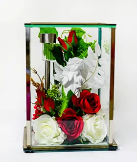 Glass and brass rectangular display case containing a white cherub statue surrounded by pink, lavender, and white roses with green foliage.