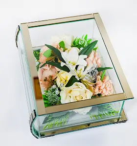 Rectangular glass and gold-trimmed display box containing an arrangement of white, cream, and peach artificial flowers with green foliage and a decorative silver butterfly.
