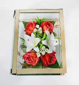 Glass rectangular display box with brass frame containing artificial flowers including white lilies, cream roses, and pink blossoms on a white surface.