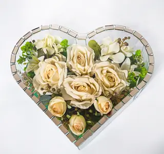 Heart-shaped glass box filled with cream roses, white flowers, and green foliage on a white background.