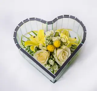 Heart-shaped glass box containing blue and white artificial flowers with green foliage.