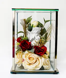 Glass rectangular display box with brass frame containing artificial flowers including white lilies, cream roses, and pink blossoms on a white surface.