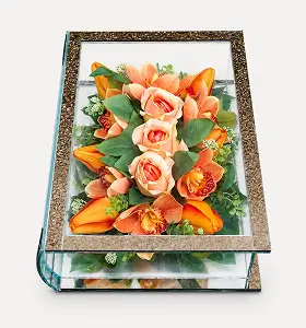 Glass rectangular display box with brass frame containing artificial flowers including white lilies, cream roses, and pink blossoms on a white surface.
