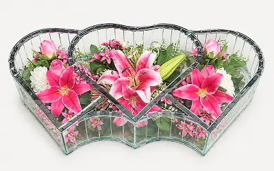 Heart-shaped glass vase containing white and peach roses with green foliage, arranged on a white surface.