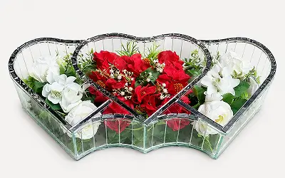 Clear three-section heart-shaped glass container holding pink roses, white flowers, and small pink blossoms on a white background.