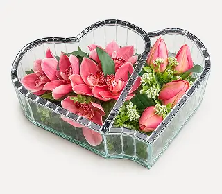 Heart-shaped mirrored tray holding peach roses, yellow flowers, two white calla lilies with decorative crystals, and a butterfly ornament.