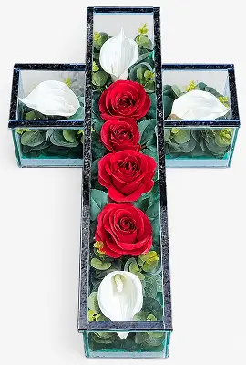 Glass cross filled with pink roses, white flowers, and green foliage on a white background.