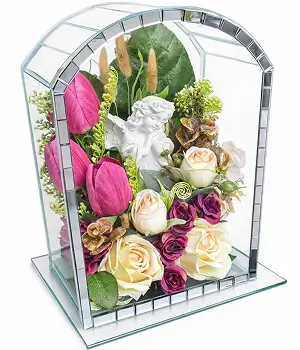 Glass rectangular display box with brass frame containing artificial flowers including white lilies, cream roses, and pink blossoms on a white surface.