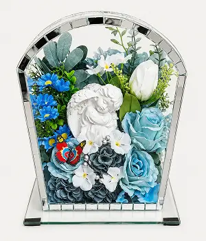 Rectangular glass and gold-trimmed display box containing an arrangement of white, cream, and peach artificial flowers with green foliage and a decorative silver butterfly.