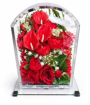 Glass rectangular display box with brass frame containing artificial flowers including white lilies, cream roses, and pink blossoms on a white surface.