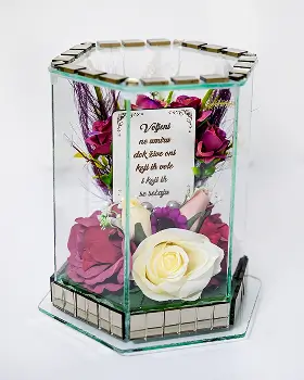 Glass hexagonal display case containing white roses, green foliage, small silver ornaments, and a white angel figurine.
