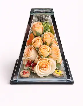 Glass pyramid display case containing a white praying Virgin Mary figurine surrounded by red roses, red daisies, and green fern leaves.