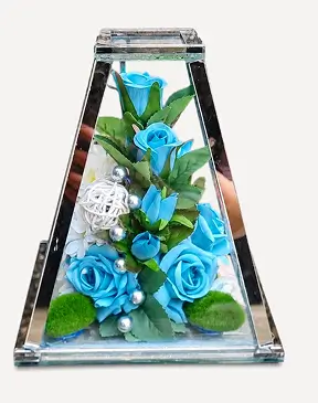 Glass pyramid frame containing white angel statue, two orange calla lilies, white roses, green leaves, and red berries on a white background.