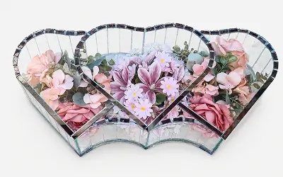 Heart-shaped mirrored glass container holding a bouquet of white and orange roses with green foliage.