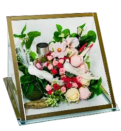 Square glass display case containing a variety of pink and white flowers with green leaves and a white angel figurine in the center.