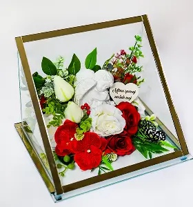 Glass box with gold trim containing a mix of red and white flowers, greenery, a small angel figurine, and a heart-shaped plaque with a message.