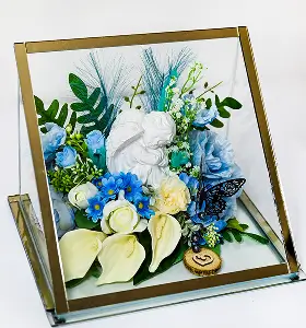 Decorative glass box containing a white cherub statue surrounded by blue and white artificial flowers and green leaves, with a small colorful butterfly.