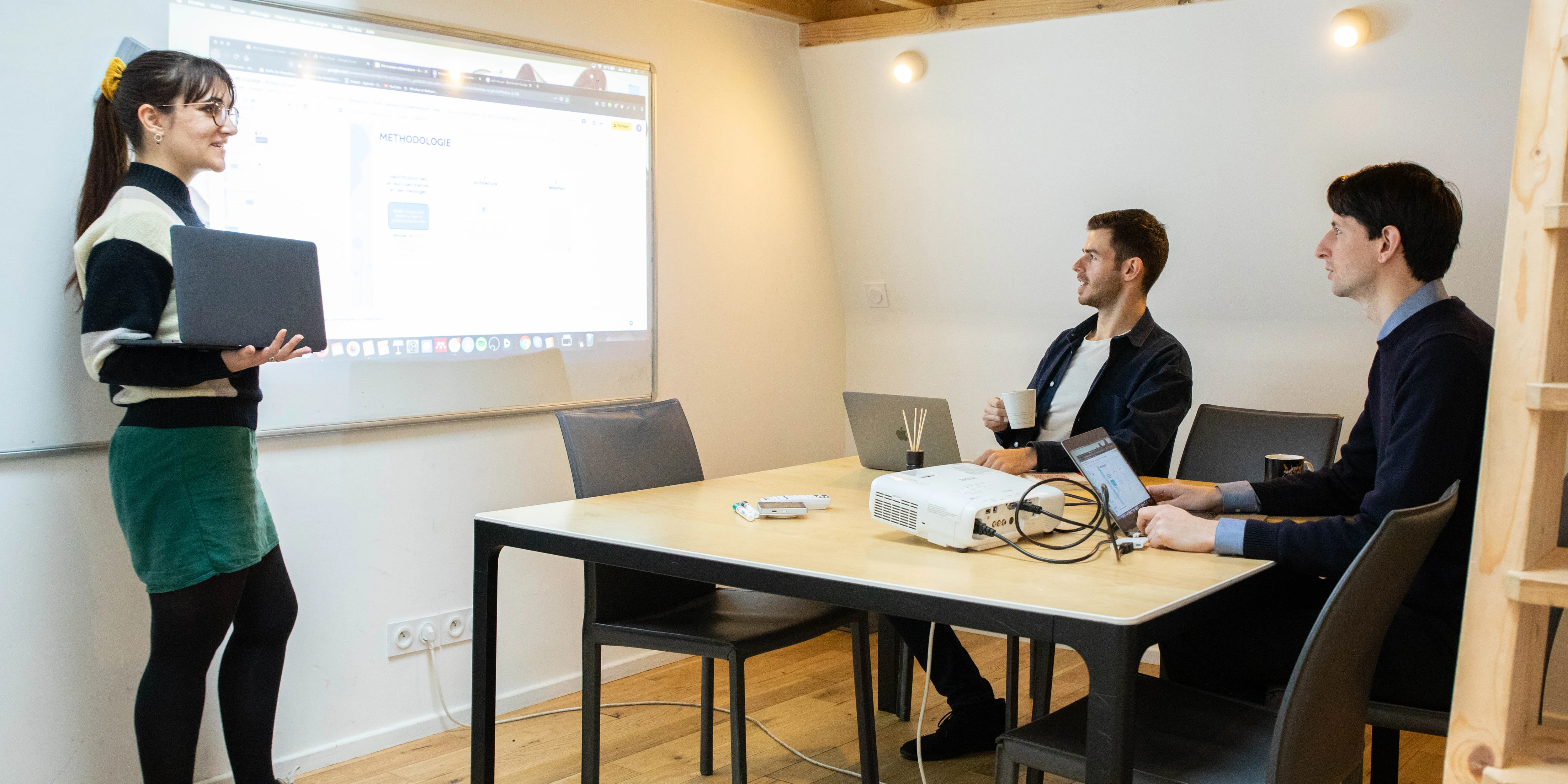 Une femme fait une présentation devant un tableau blanc projeté, tandis que deux hommes écoutent assis à une table avec des ordinateurs portables.