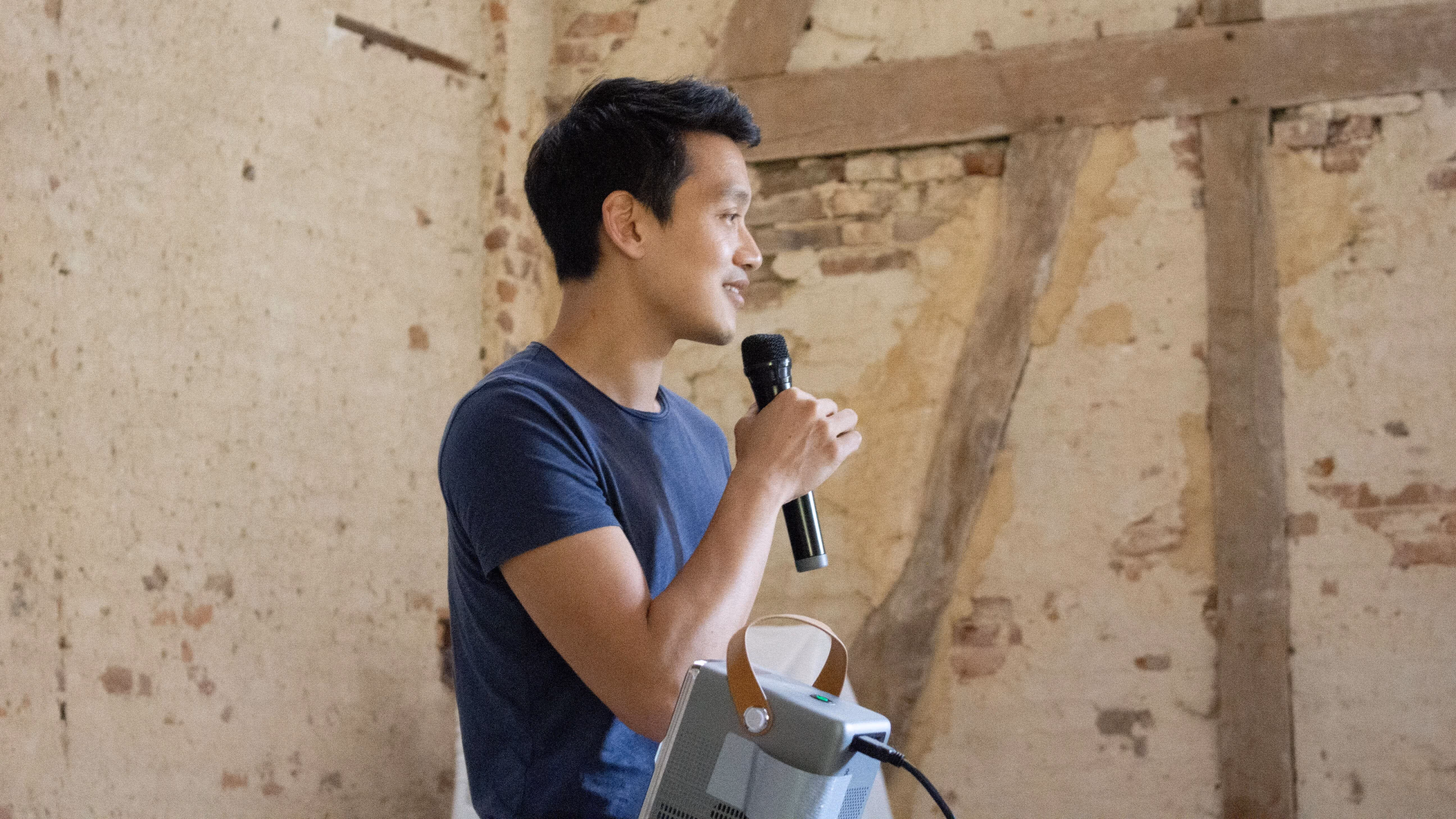 Son Ly souriant tenant un microphone, debout devant un mur en briques avec des poutres en bois apparentes.