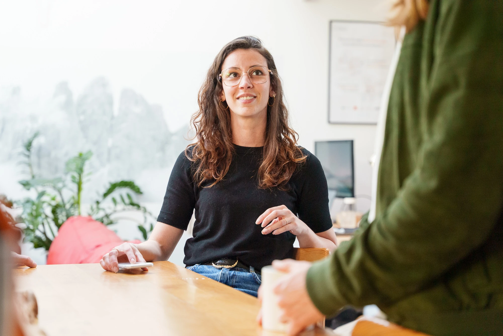 Jeune femme aux cheveux longs et lunettes souriant, assise à une table en bois, en conversation avec une personne en veste verte.