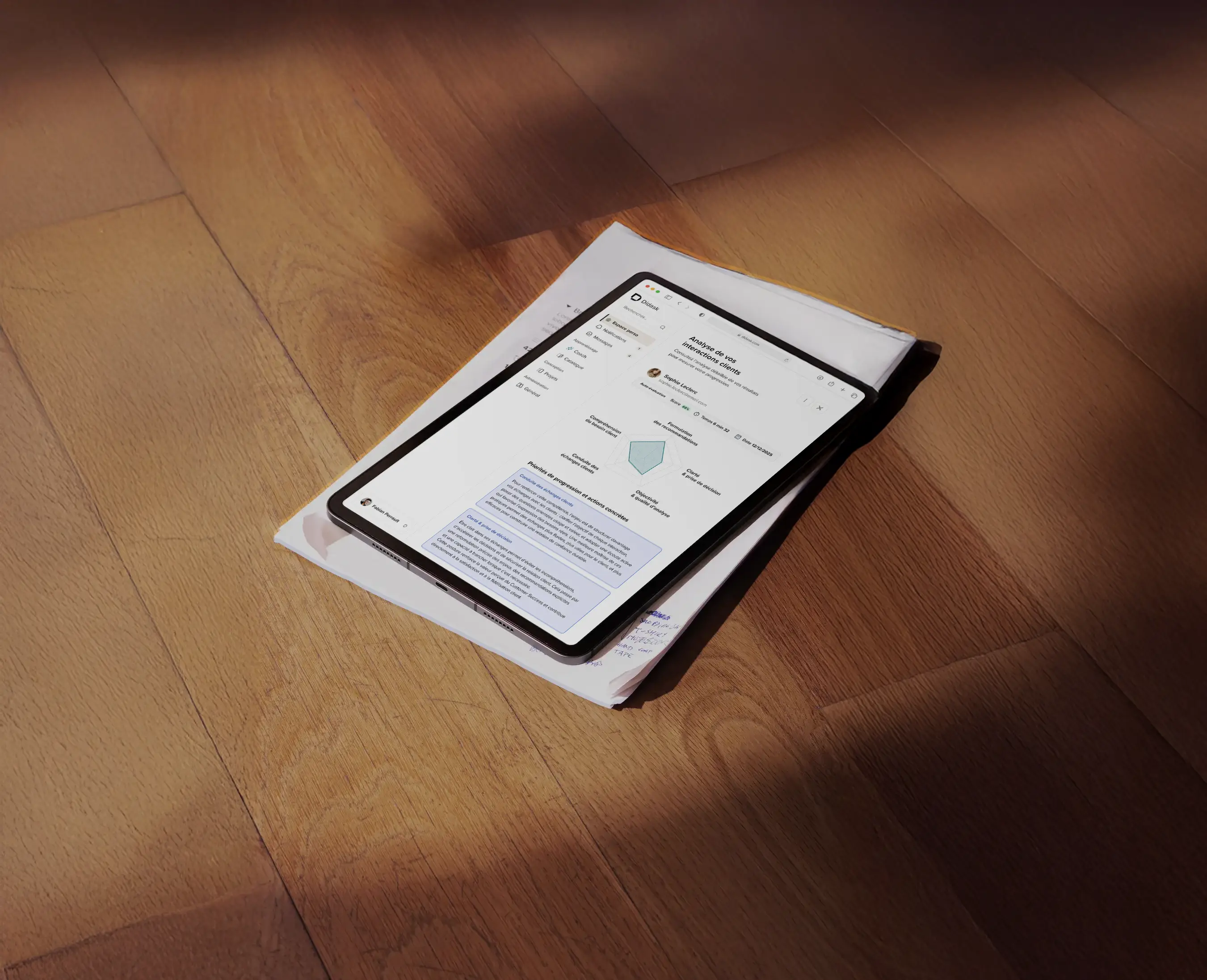Tablette posée sur un tas de papiers sur un sol en bois, affichant une interface d'analyse des interactions clients en français.