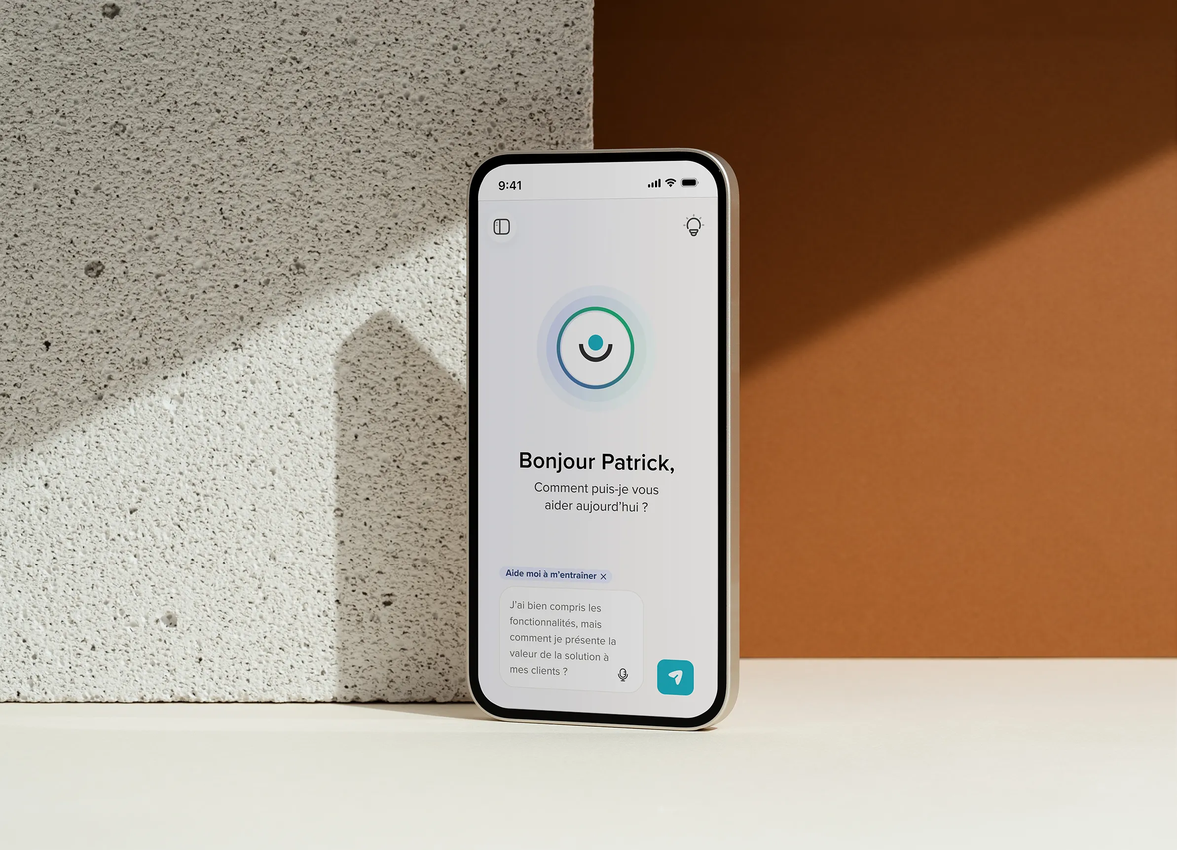 Un smartphone posé sur une surface en bois affiche une application avec le message « Bonjour Patrick, comment puis-je vous aider aujourd'hui ? » et une question en bas.