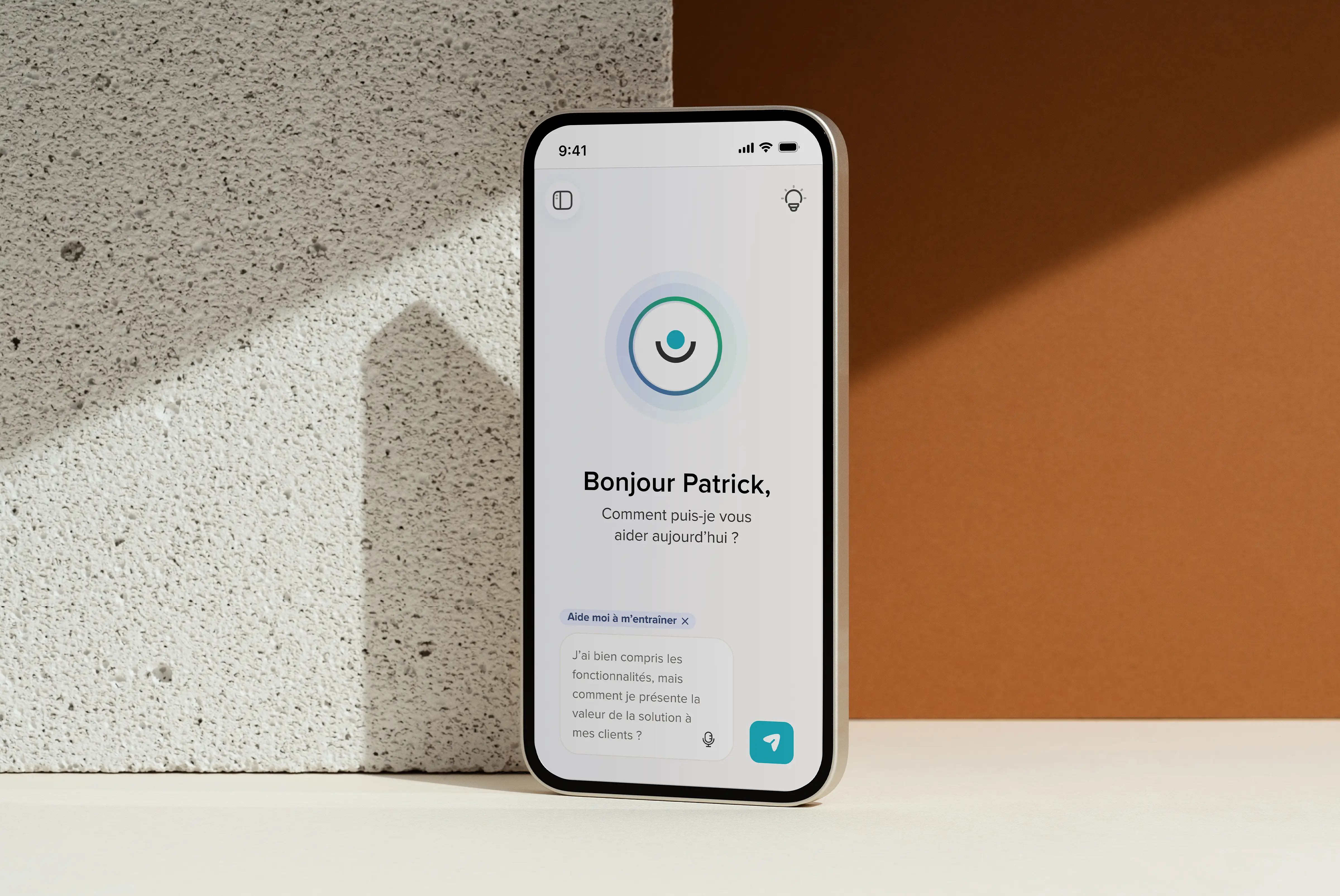 Un smartphone posé sur une surface en bois affiche une application avec le message « Bonjour Patrick, comment puis-je vous aider aujourd'hui ? » et une question en bas.