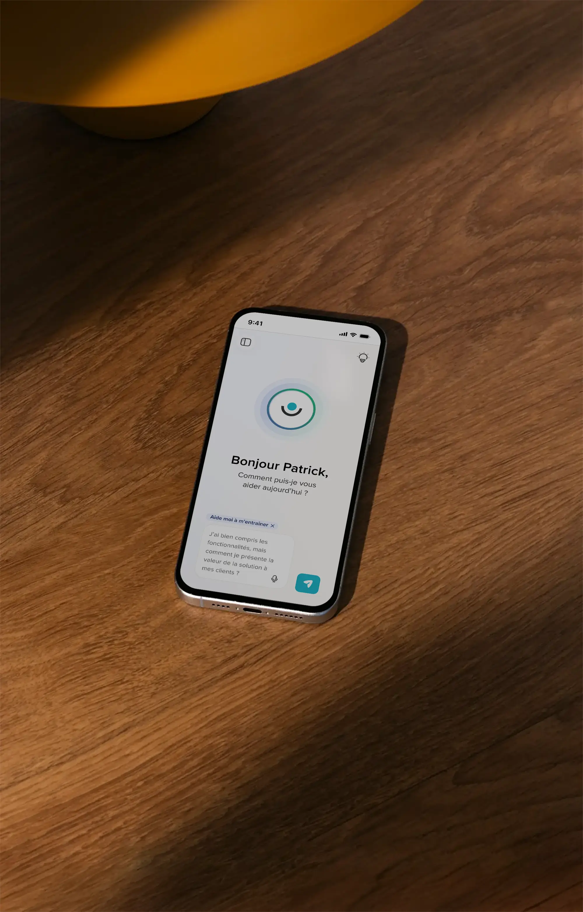 Un smartphone posé sur une surface en bois affiche une application avec le message « Bonjour Patrick, comment puis-je vous aider aujourd'hui ? » et une question en bas.