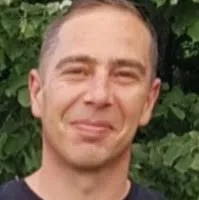 Portrait d'un homme souriant aux cheveux courts devant un feuillage vert.