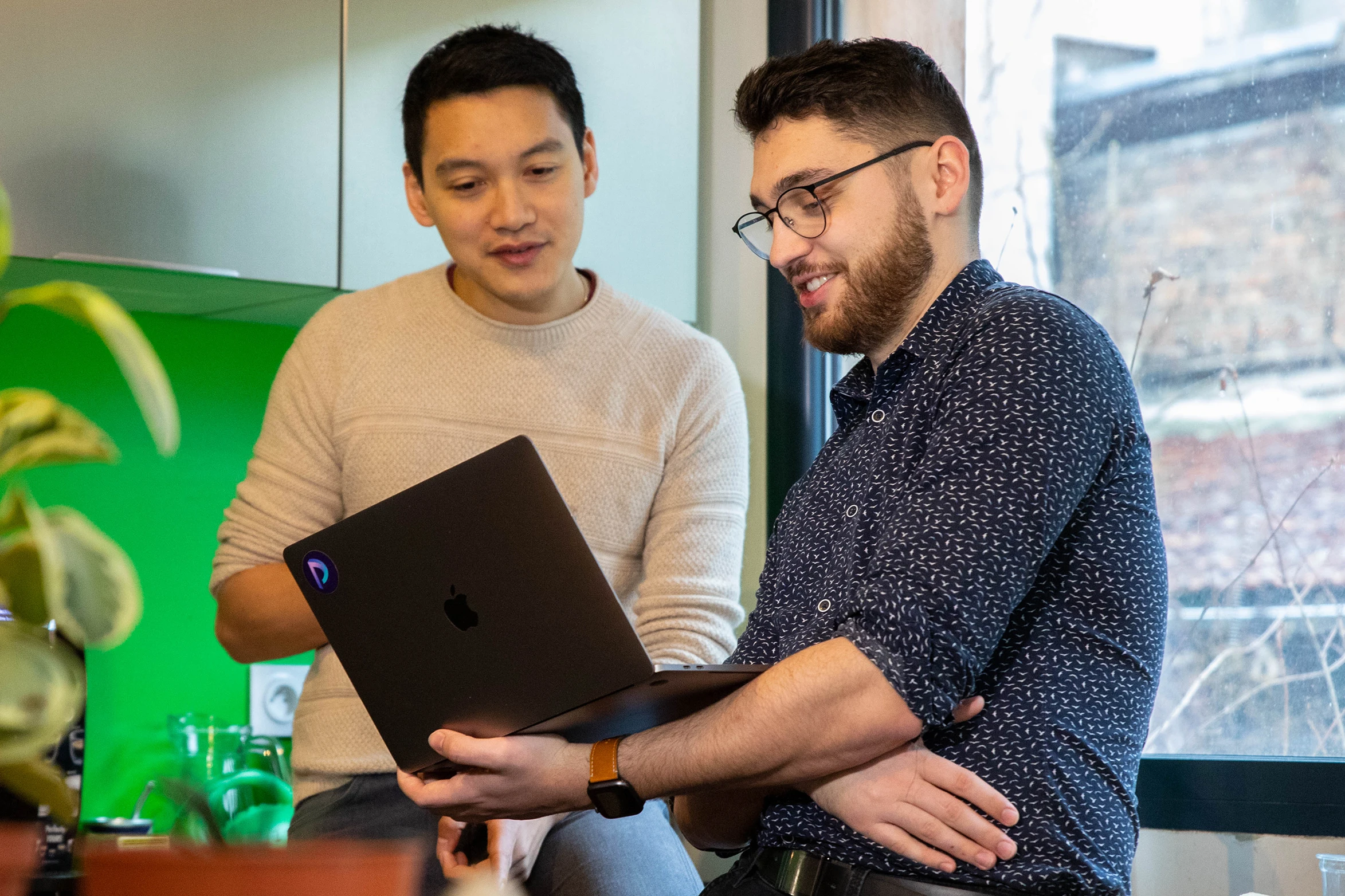 Deux hommes discutant en regardant un ordinateur portable dans un intérieur avec une fenêtre.