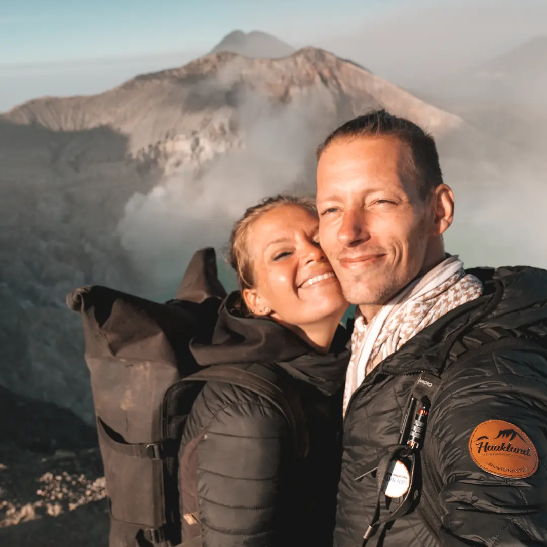Ein Paar in Wanderbekleidung posiert glücklich für ein Selfie vor nebligen Vulkanbergen.